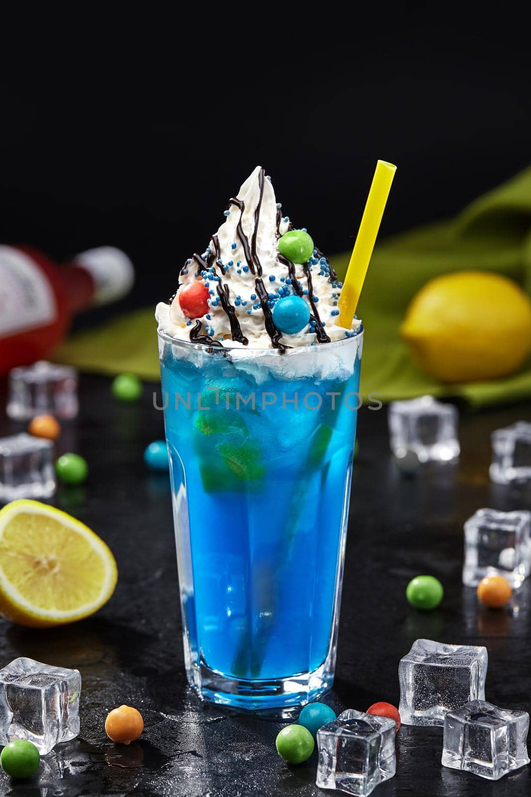 Freaky blue lemonade with cap of delicate whipped cream topped with chocolate icing, colorful sprinkle and balls of chewing gum on black background. Unusual mix of refreshing drink and sweet dessert