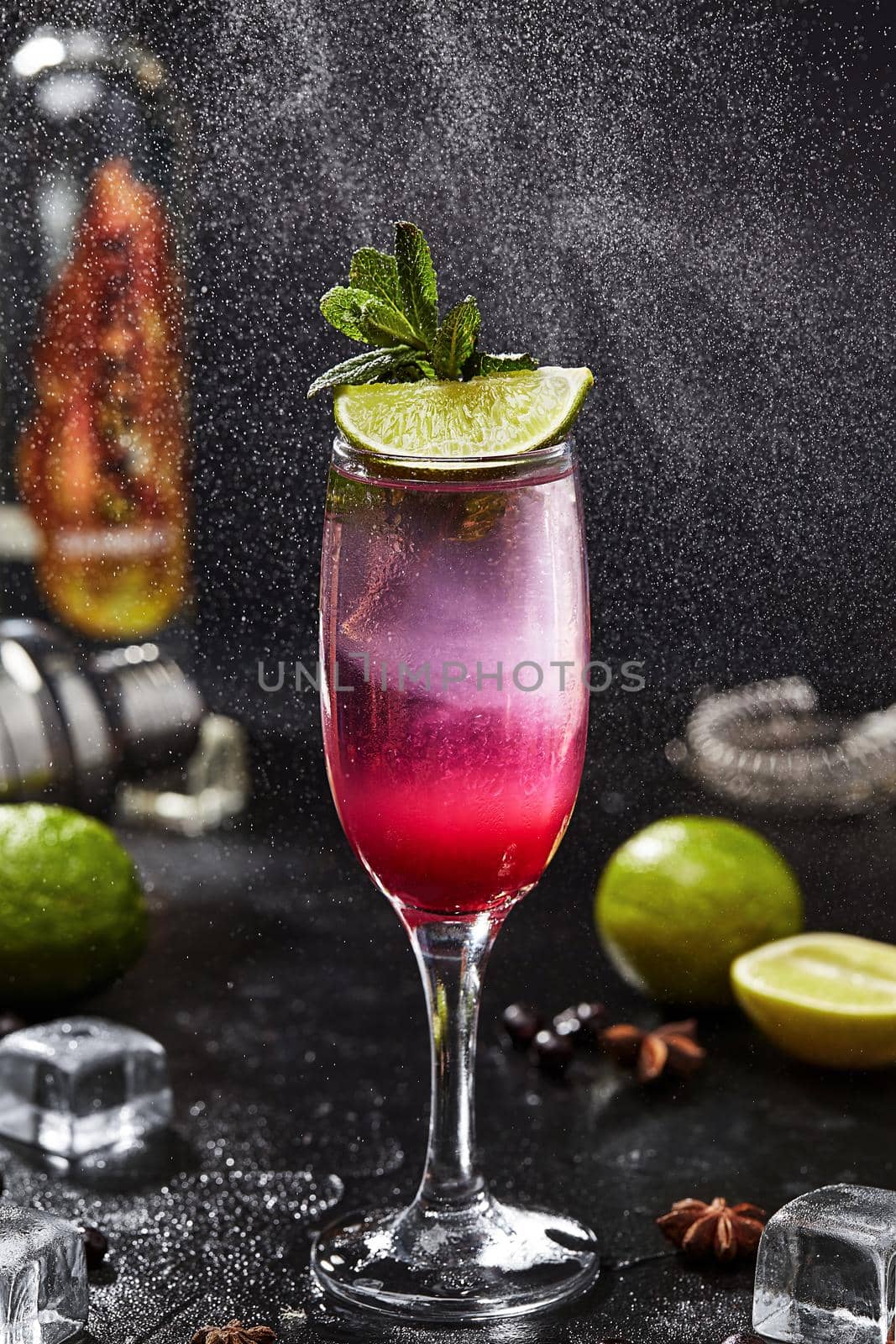Refreshing alcoholic cocktail based on sambuca and white rum with lime juice, soda and currant syrup served in glass garnished with lime slice and mint leaves on black background in water spray