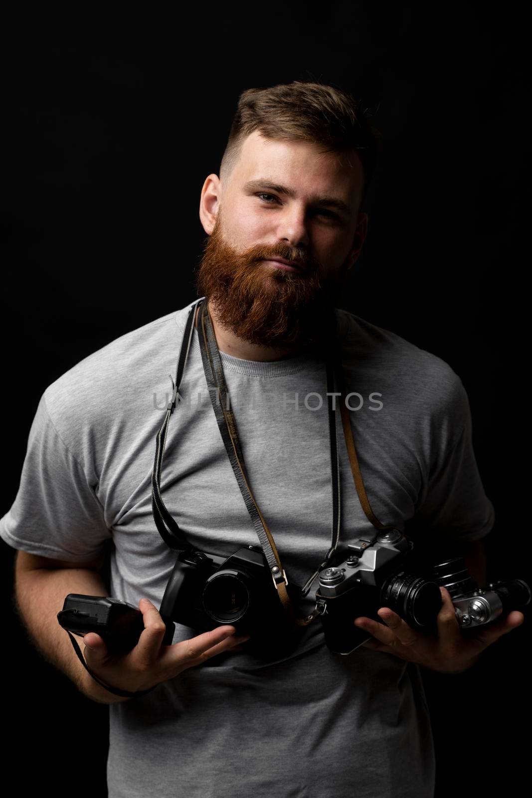 Portraite of bearded professional photographer in a grey t-shirt with a bunch of different cameras in a hands and on a shoulder looking on a camera and ready for make a good shoot. by vovsht