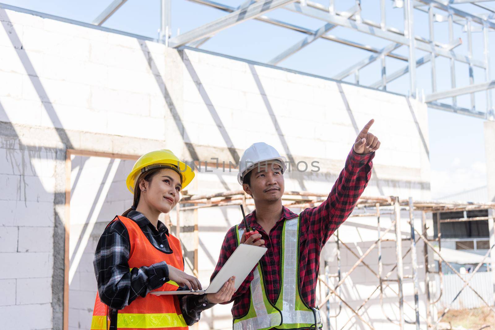 Asian engineer foreman worker man and woman working at building construction site use laptop and talking with radio, engineering hold computer and radio discuss and control worker employee to building