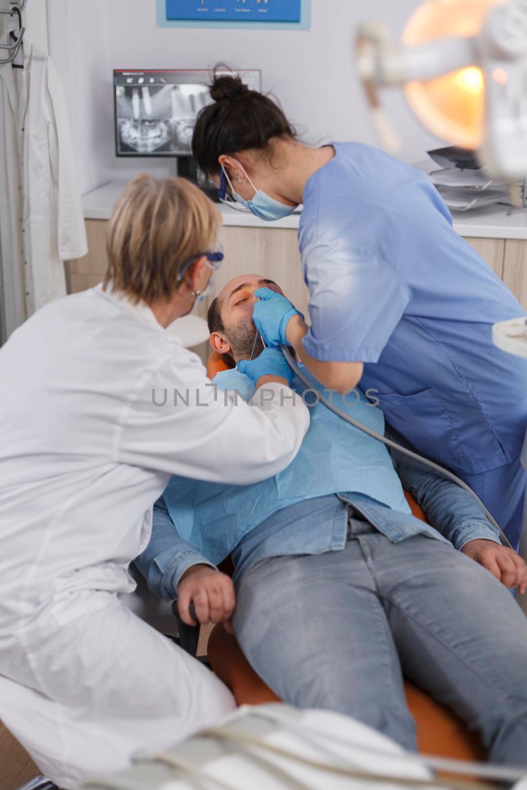 Specialist dentist team with face mask examining patient mouth during medical surgery in stomatological office room. Senior woman doctor using professional dentistry tools for cavity procedure