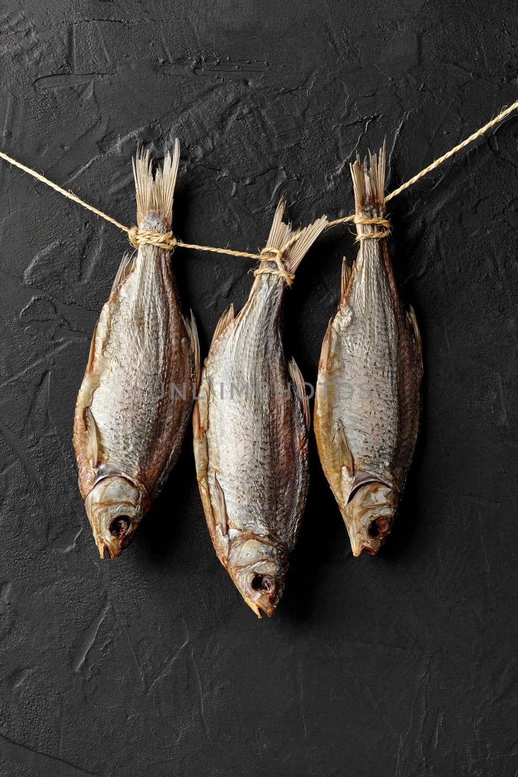 Three dried salted roach fish hanging by tails on rope on black wall