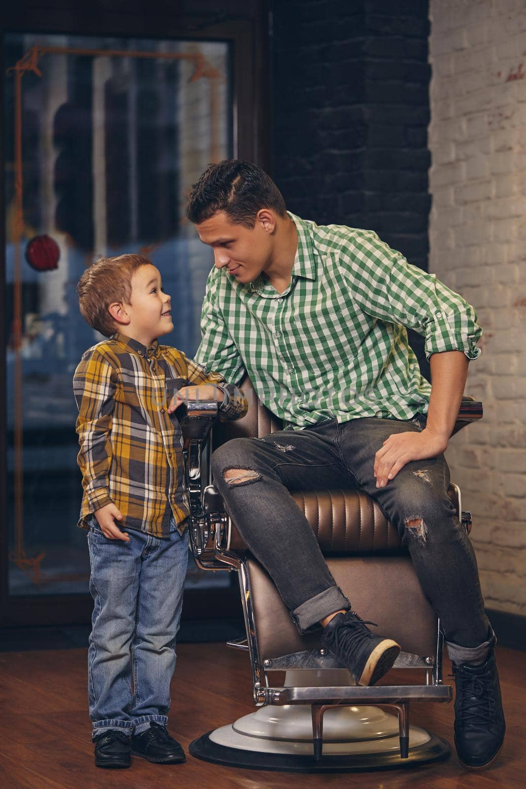 Young handsome father and his little stylish son at barbershop waiting for barber. The father is sitting in the chair and the son is standing nearby