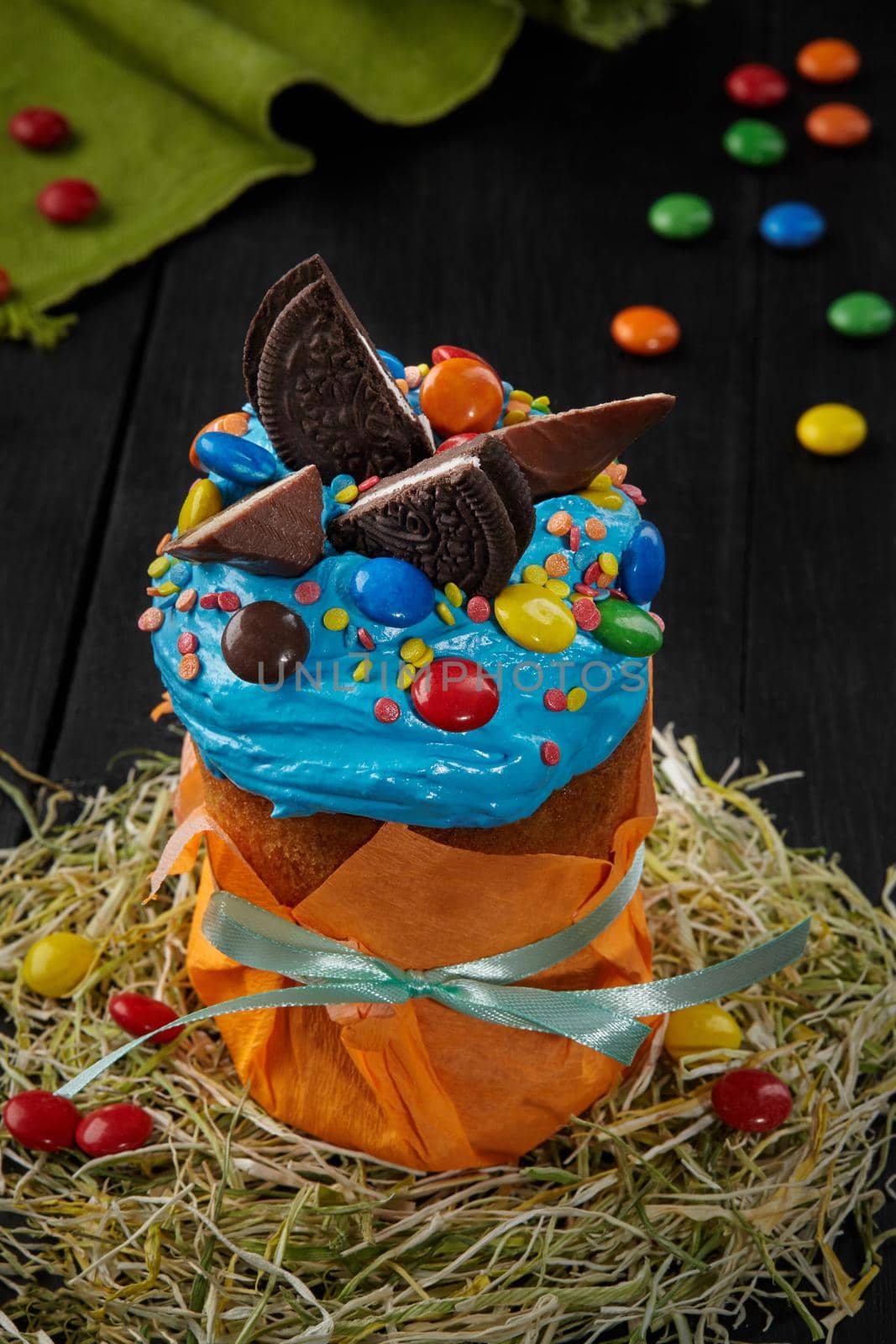 Delicious Easter cake in orange paper wrapping decorated with blue whipped egg whites icing, colorful sprinkles and dragee, chocolate cookies and candy bar slices standing on hay on black wooden table