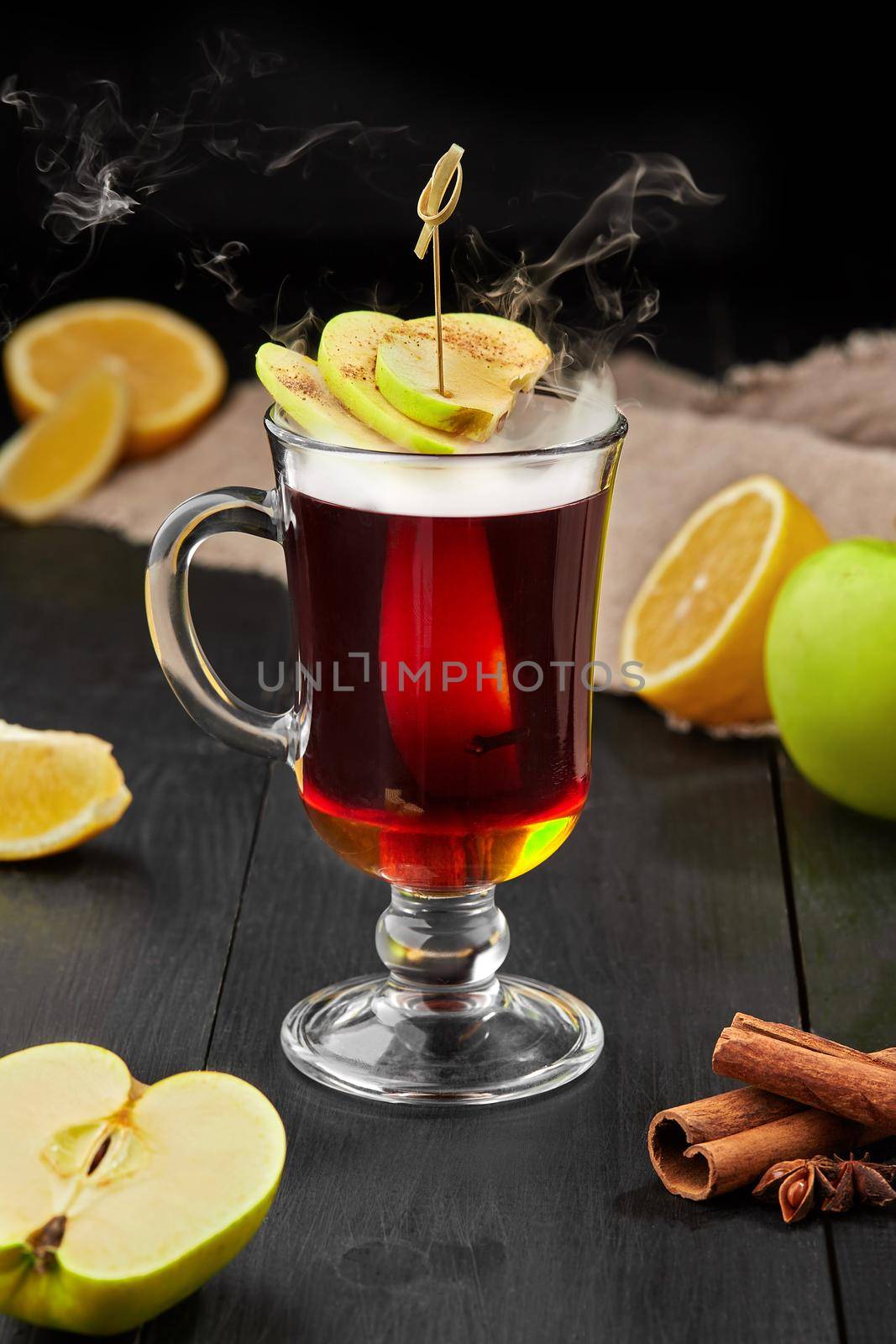 Glass cup with warming aromatic winter tea with fresh apples, lemon slices and fragrant cinnamon, cloves and star anise on black wooden table. Healthy vitamin drinks concept