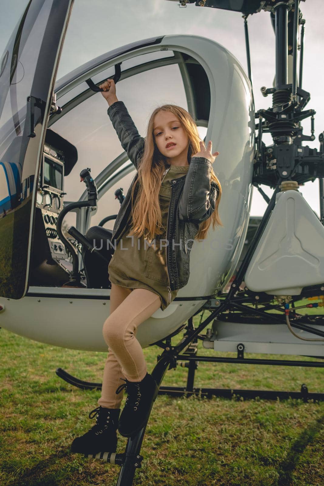 Cute brown-haired preteen girl wearing black leather jacket standing on footboard of open helicopter landed on green flying field