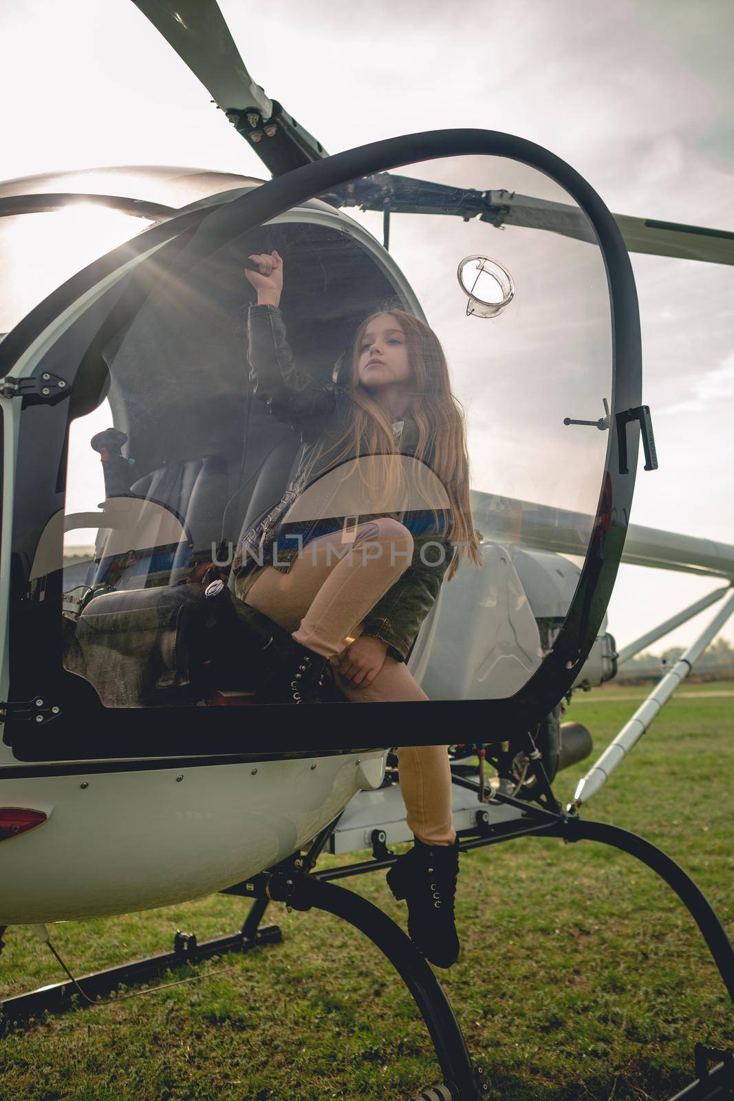 Preteen girl sitting in cockpit of landed modern helicopter in pilots seat behind glass of open door in rays of autumn sun breaking through clouds