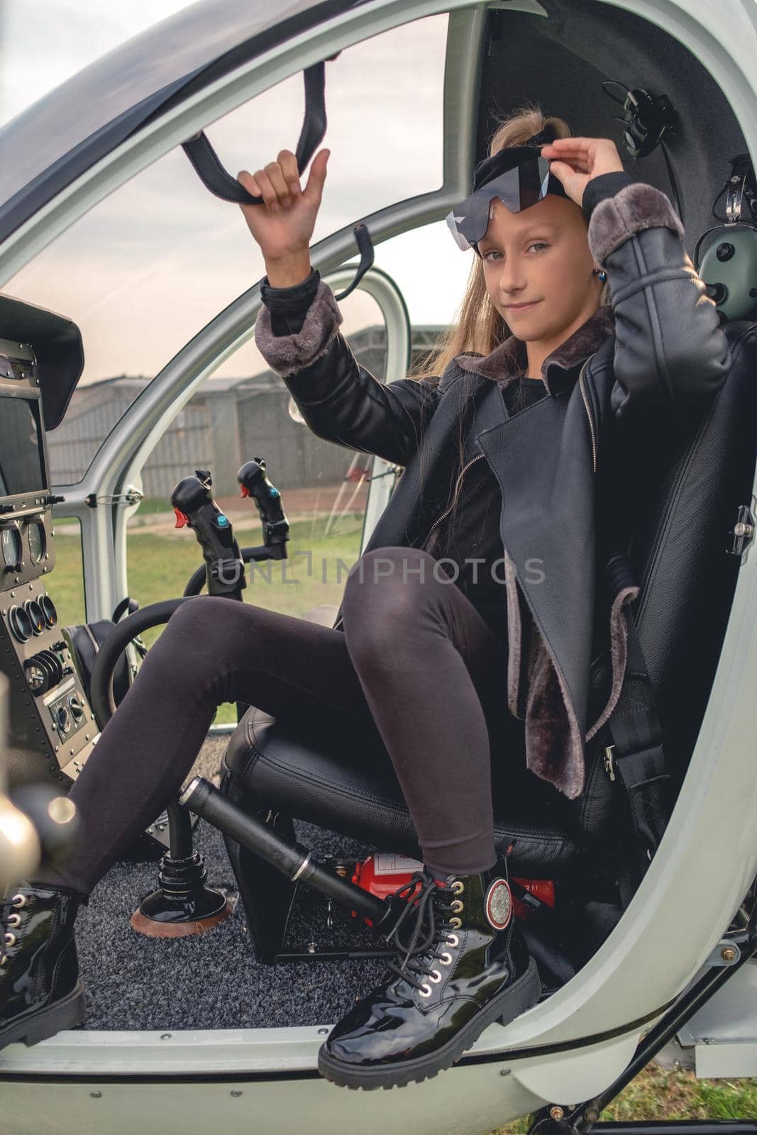 Long-haired tween girl looking confidently at camera lifting slightly her sunglasses while sitting in cockpit of light passenger helicopter on spring day