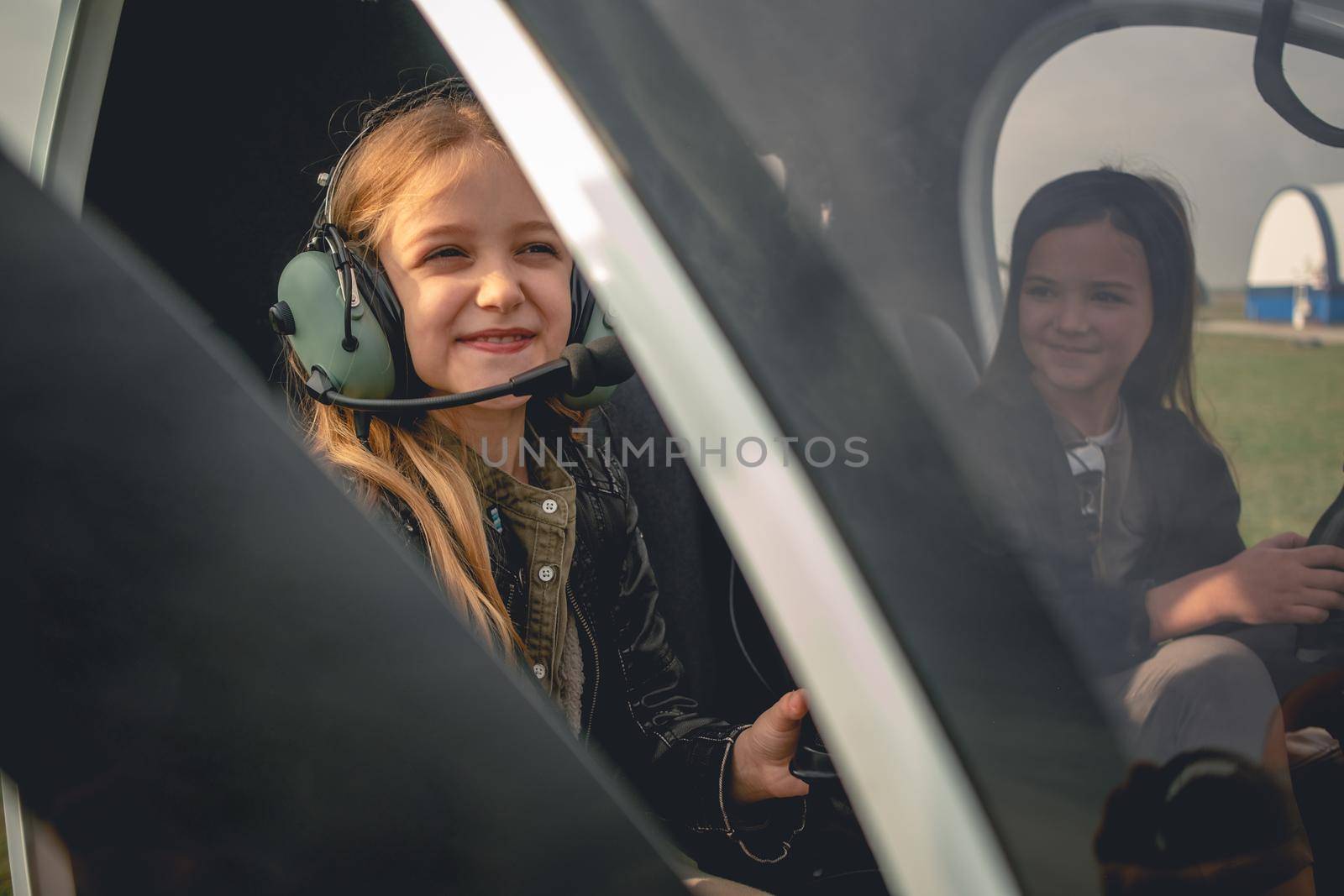 Portrait of cute smiling tween girl wearing pilot headset sitting with her female friend in helicopter landed at airfield. Happy childhood and dreams implementation concept