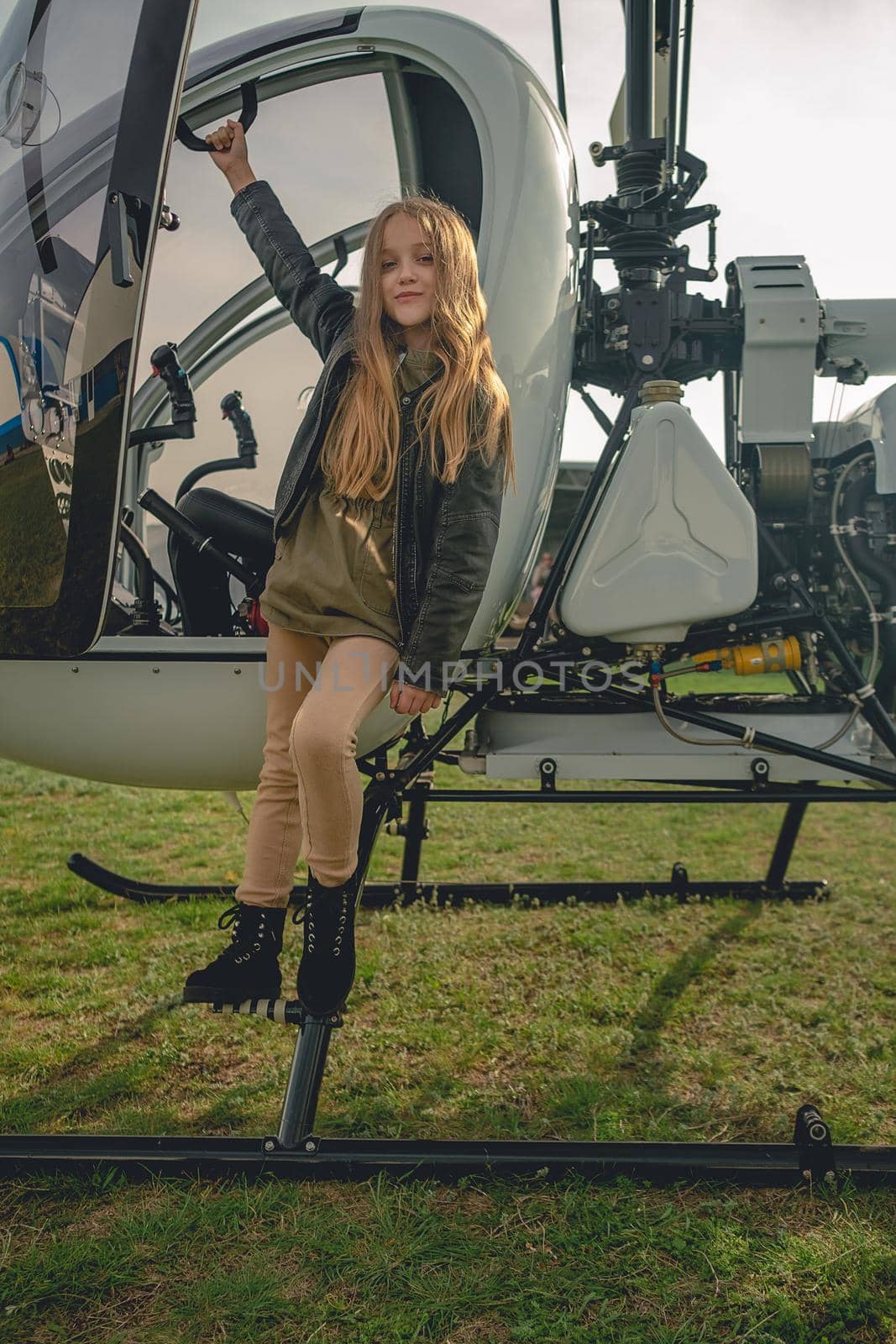 Portrait of cute brown-haired preteen girl standing on footboard of open helicopter landed on green flying field