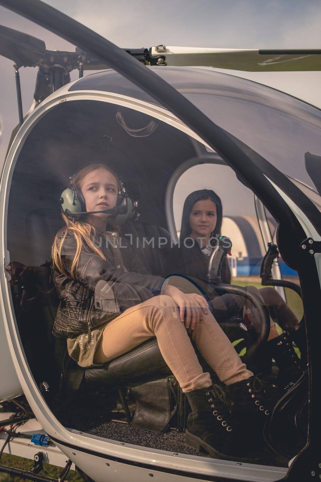 Two dreamy tween girls sitting on pilot seats in cockpit of modern helicopter landed at flying field, looking into distance