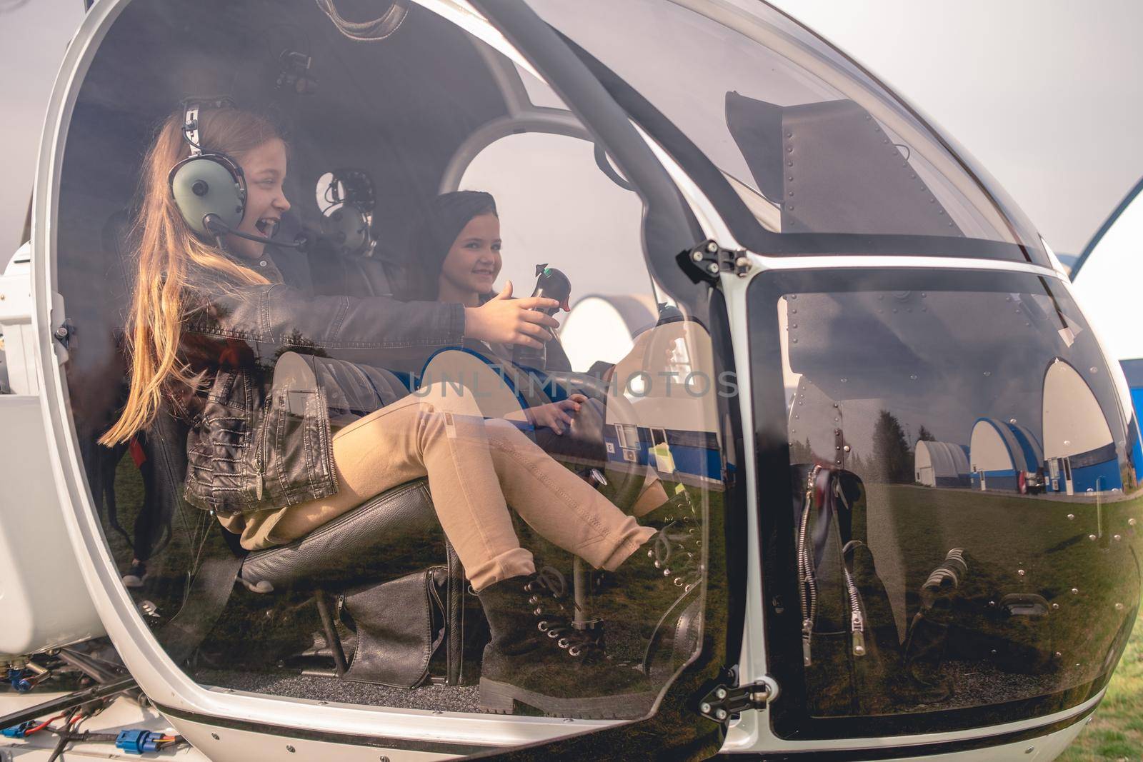 Happy emotional tween girl wearing professional aviator headphones sitting on co-pilot seat in open cockpit with her friend, holding control stick and simulating helicopter control