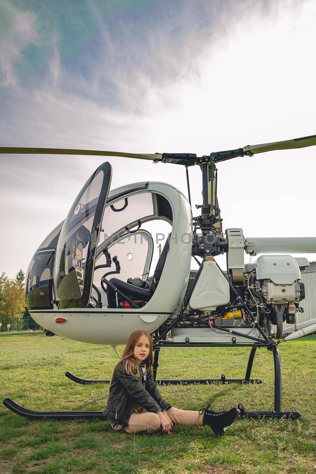 Long-haired tween girl sitting on green grass of flying field near open cockpit of modern white helicopter on spring day