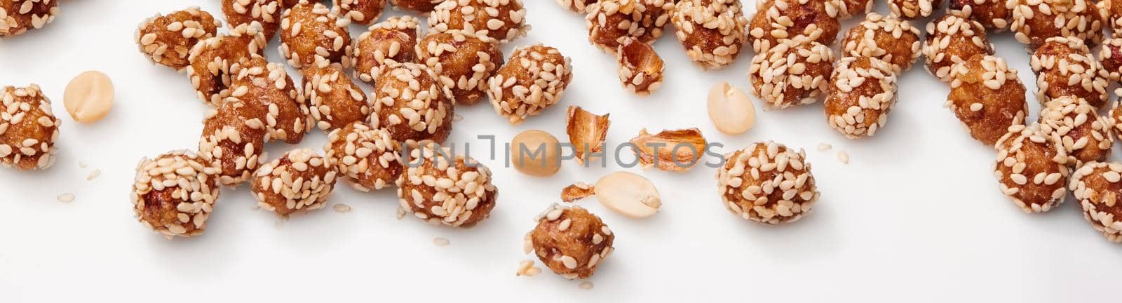 Closeup of delicious candied peanuts in honey and sugar glaze, sprinkled with sesame scattered on white surface. Concept of sweet cracker nuts as snacks. Horizontal image of natural food background