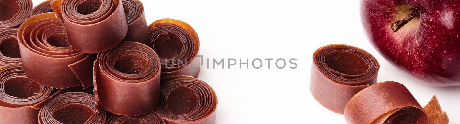 Closeup of rolls of fruit leather stacked on white surface with fresh ripe red apple. Concept of popular fruit sweets and organic snacks. Natural food background with copyspace