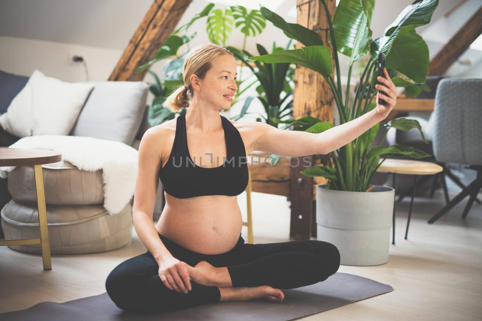 Young happy and cheerful beautiful pregnant woman taking selfie with her mobile phone while staying fit, sporty and active on her maternity leave. Motherhood, pregnancy, yoga concept. by kasto