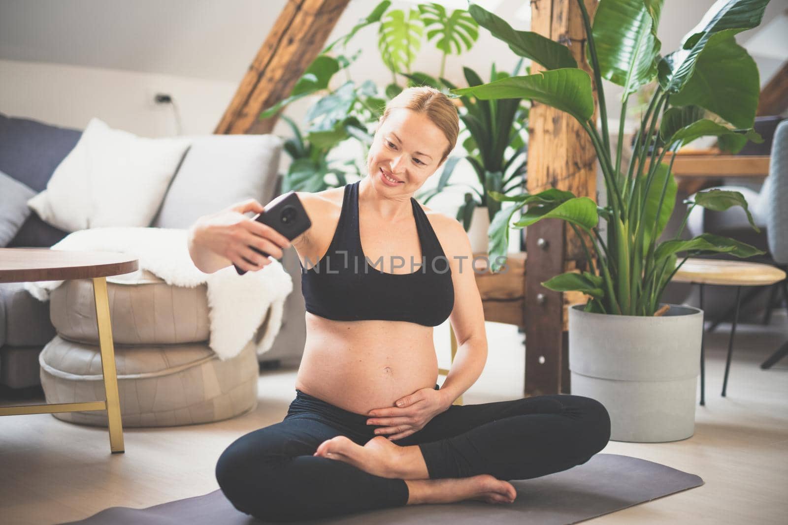 Young happy and cheerful beautiful pregnant woman taking selfie with her mobile phone while staying fit, sporty and active on her maternity leave. Motherhood, pregnancy, yoga concept. by kasto