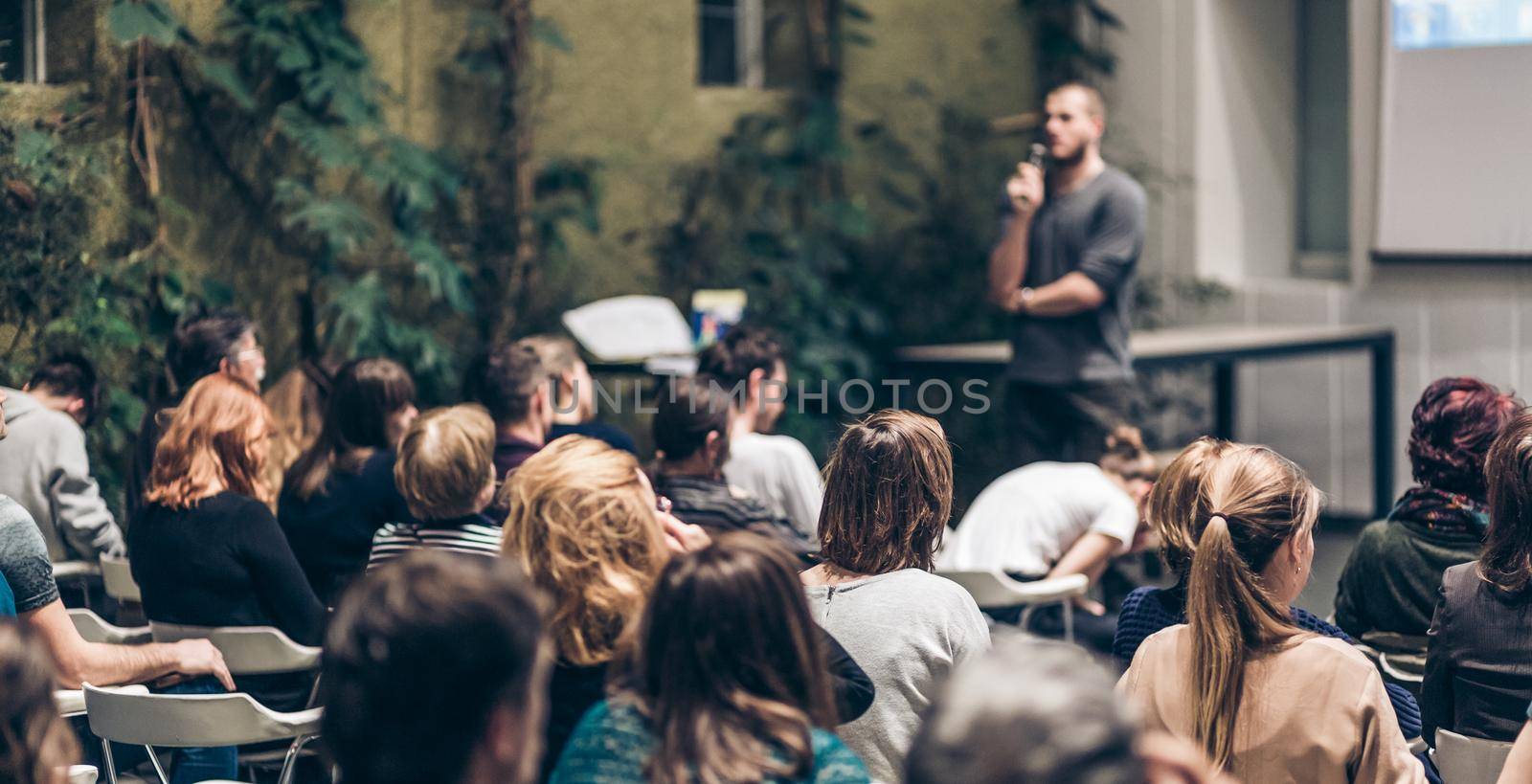 Male speaker giving presentation in lecture hall at university workshop. Audience in conference hall. Rear view of unrecognized participant in audience. Scientific conference event.