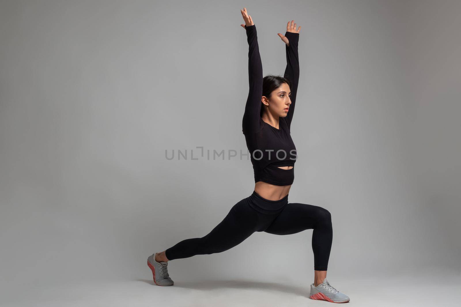 Girl doing forward lunges with arms stretched upwards on grey background by nazarovsergey