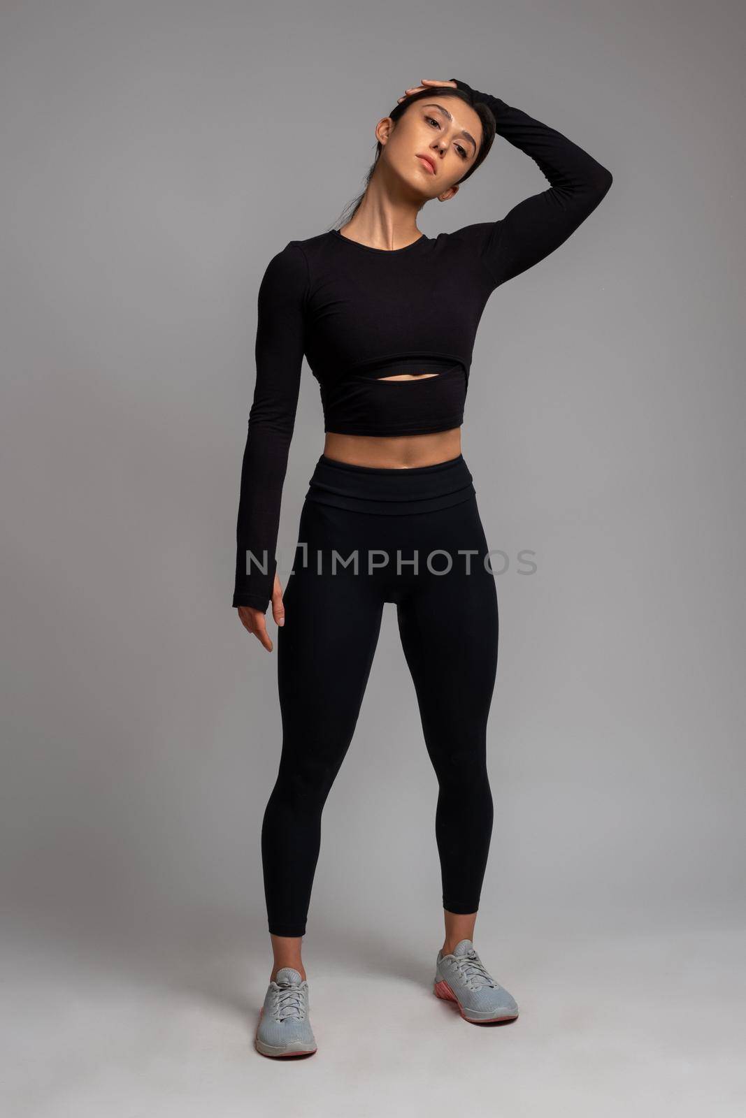 Confident young sportswoman warming up before intense workout, doing stretching exercises. Full length studio portrait on grey background