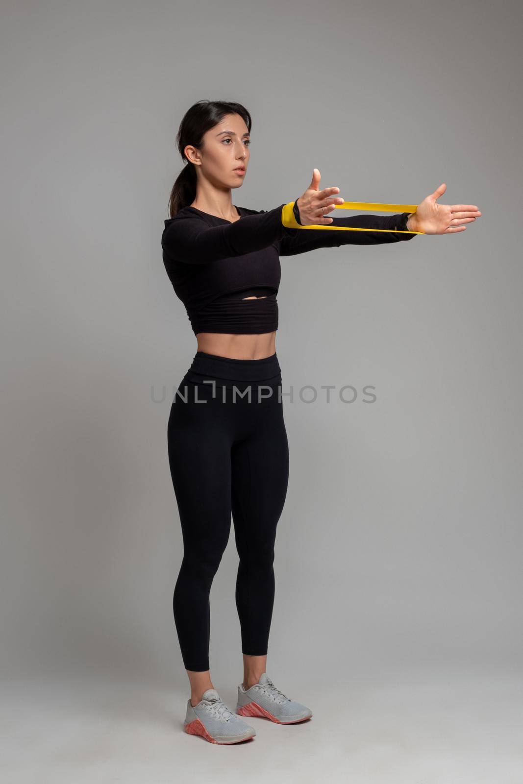 Athletic girl in black sportswear doing exercise to sculpt and tone arms, stretching resistance elastic band on outstretched arms on grey background. Sport and fitness concept