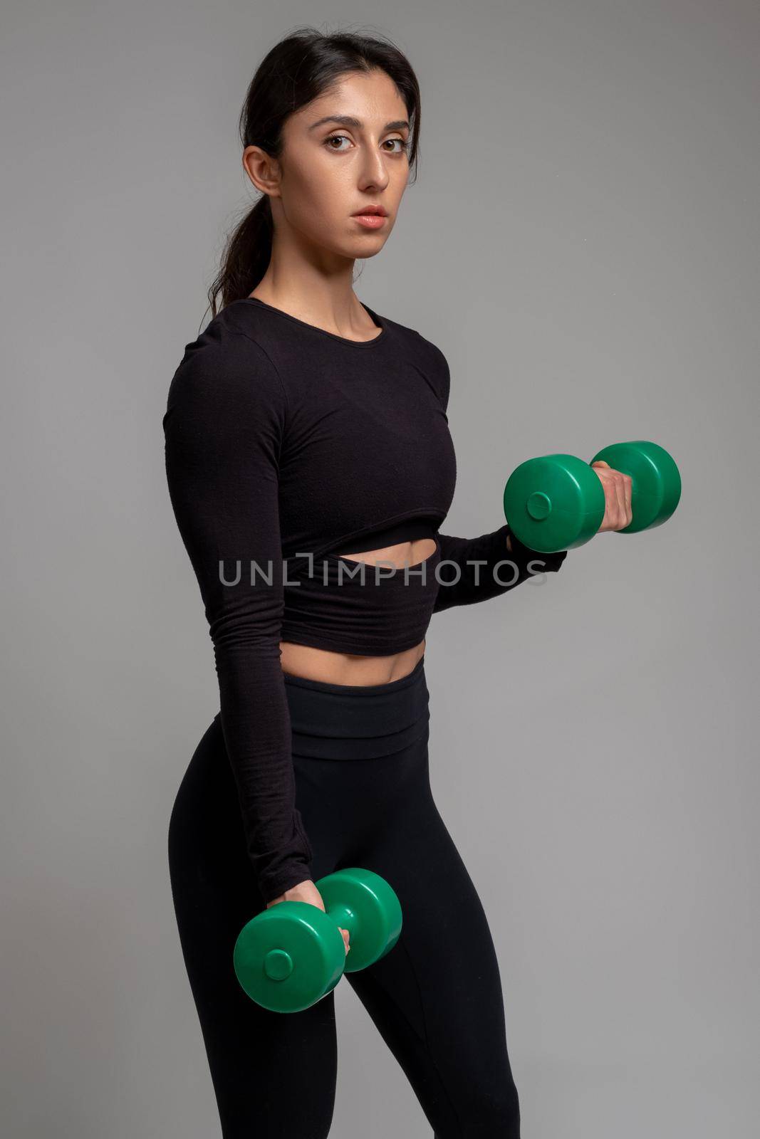 Athletic girl performing set of arm exercises with dumbbells in studio on grey background, confidently looking at camera. Sports motivation concept