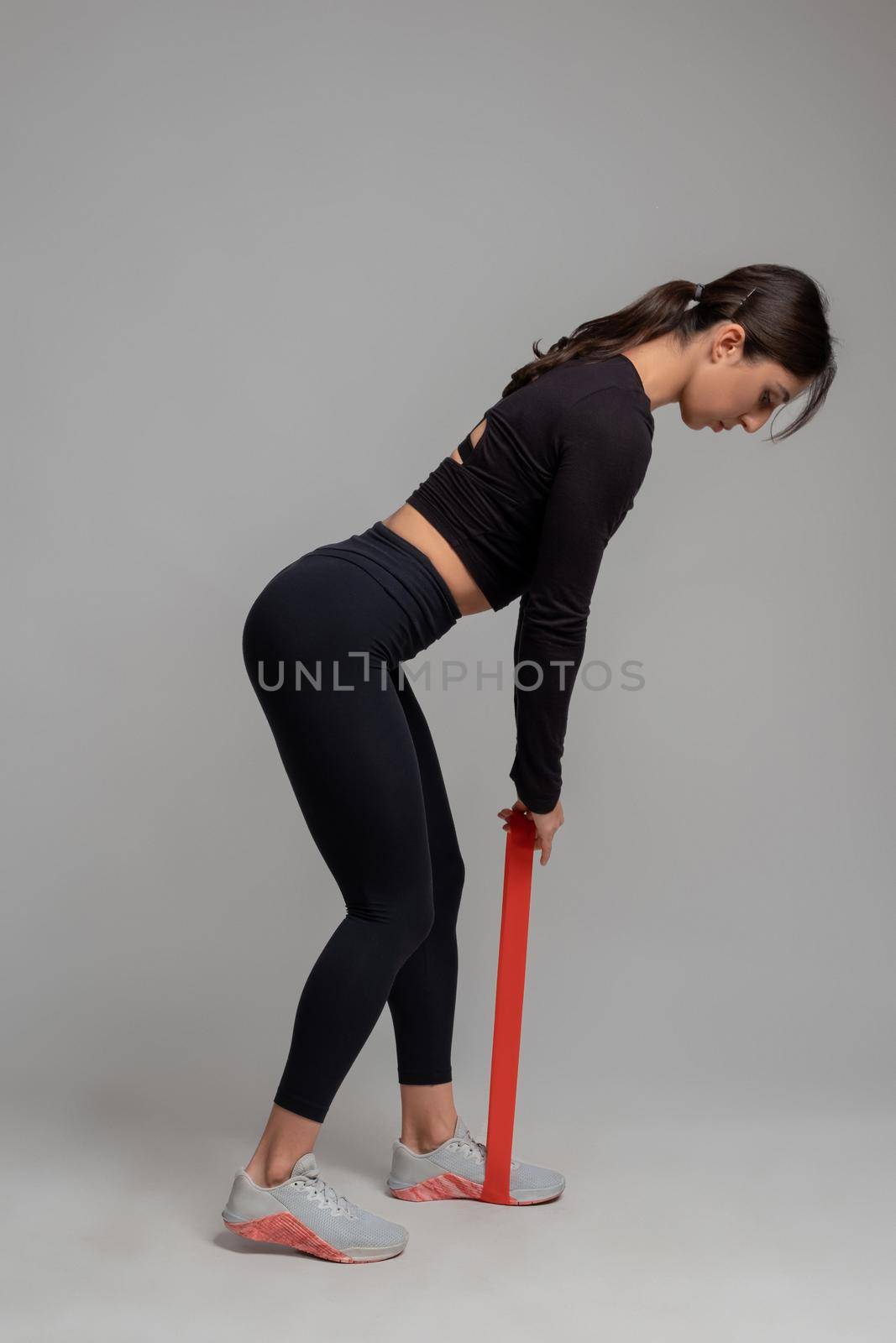 Sporty girl working out muscles of legs and glutes, doing Romanian resistance loop deadlift in studio on grey background. Fitness and sports motivation concept