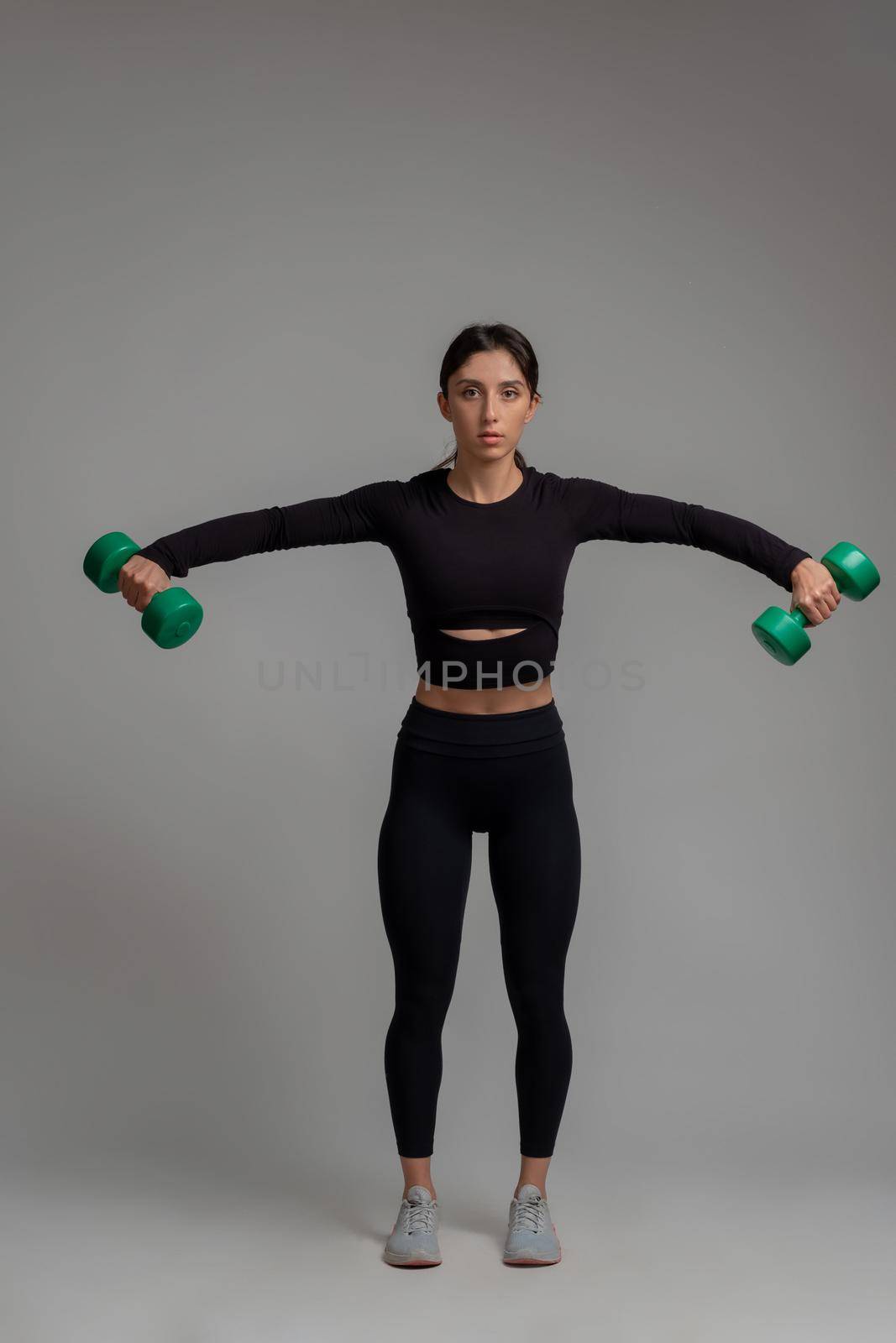 Sporty girl in black activewear working out shoulders muscles in studio on grey background, performing dumbbell lateral raise. Physical activity and fitness concept