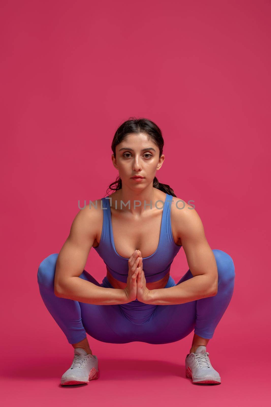 Concentrated sporty young woman doing stretching squats before intense workout on maroon background. Fitness and body conscious concept