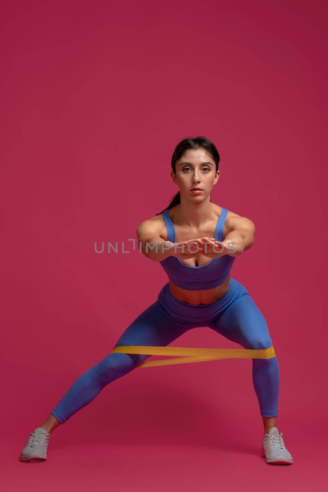 Sporty girl working out muscles of legs and glutes, doing lateral lunges with resistance loop band in studio on maroon background. Fitness and sports motivation concept