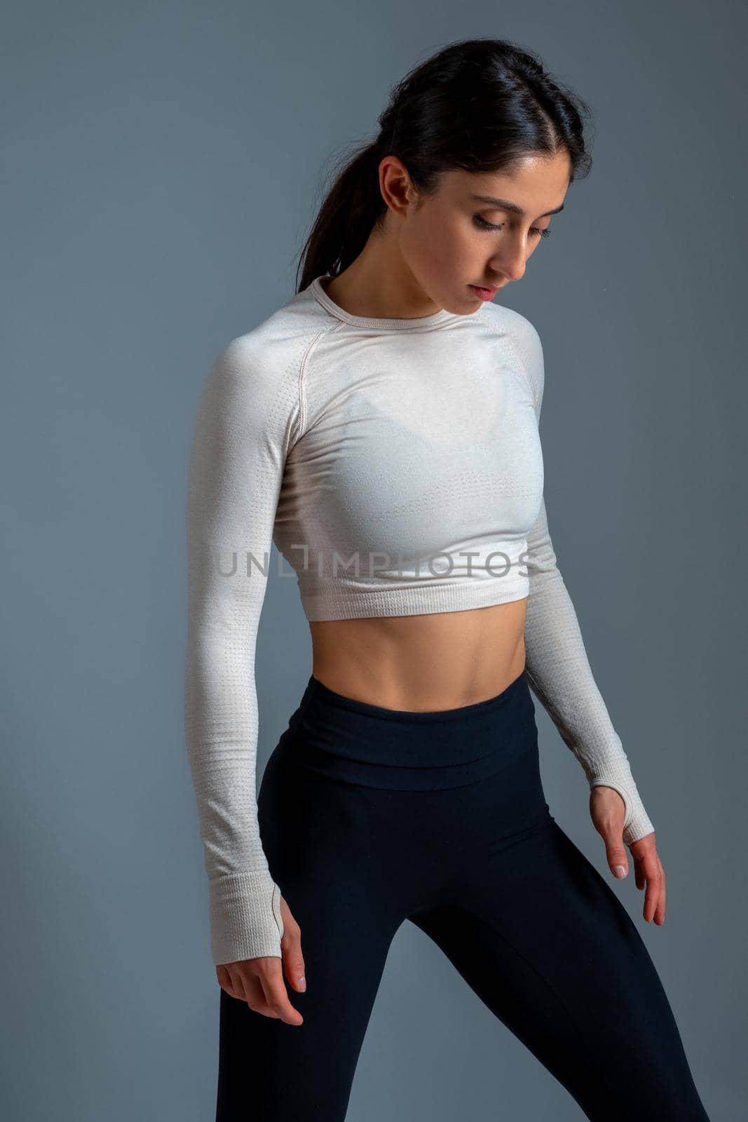 Portrait of relaxed athletic young brunette wearing sporty long sleeve crop top and leggings standing in studio interior on grey background. Sport training concept