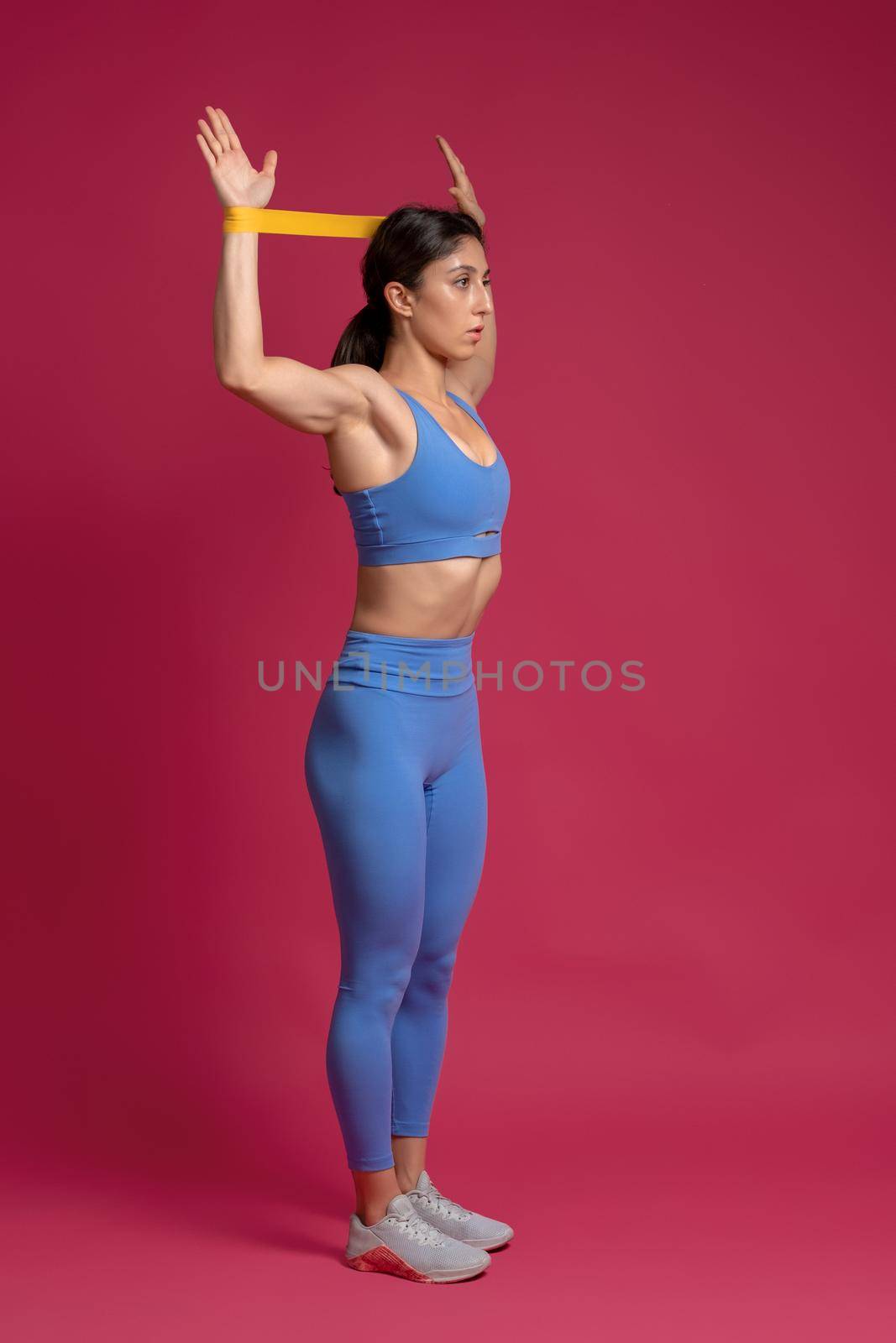 Full length portrait of athletic girl performing set of exercises for arms, shoulder and back with resistance band loop on maroon background. Sports motivation concept