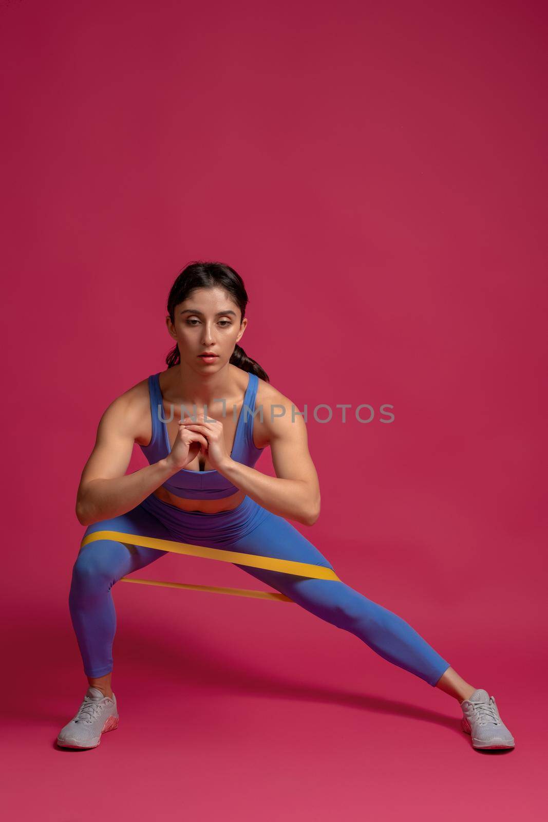 Athletic girl working out in studio interior on maroon background, doing lateral lunge to right with resistance loop band. Leg and glute workout. Active lifestyle and fitness concept