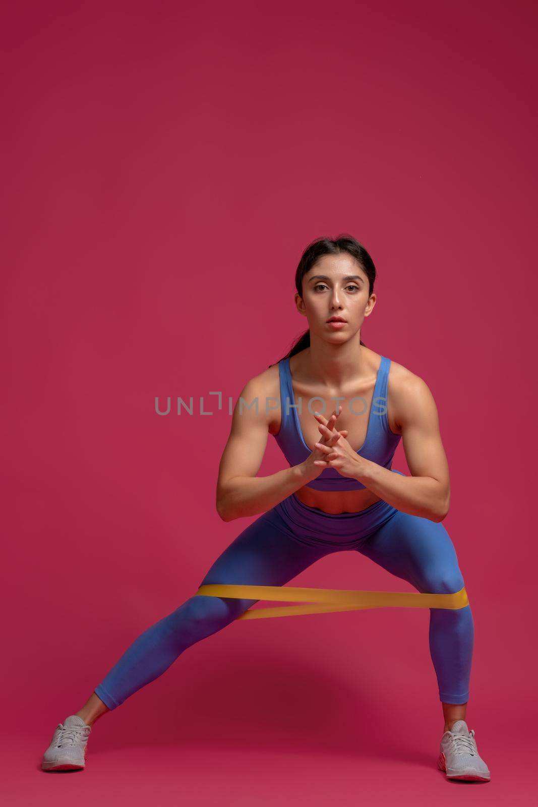 Attractive athletic girl dressed in blue sports top and leggings doing side lunges with resistance elastic band on maroon background. Concept of weight training for woman