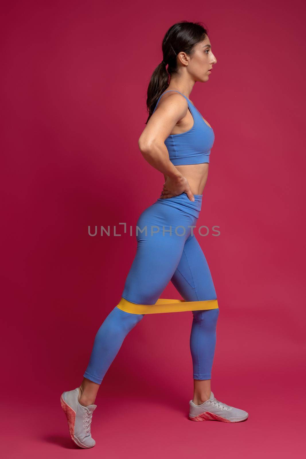 Athletic young woman performing leg and glutes training with loop resistance band in studio on maroon background. Active lifestyle and fitness concept