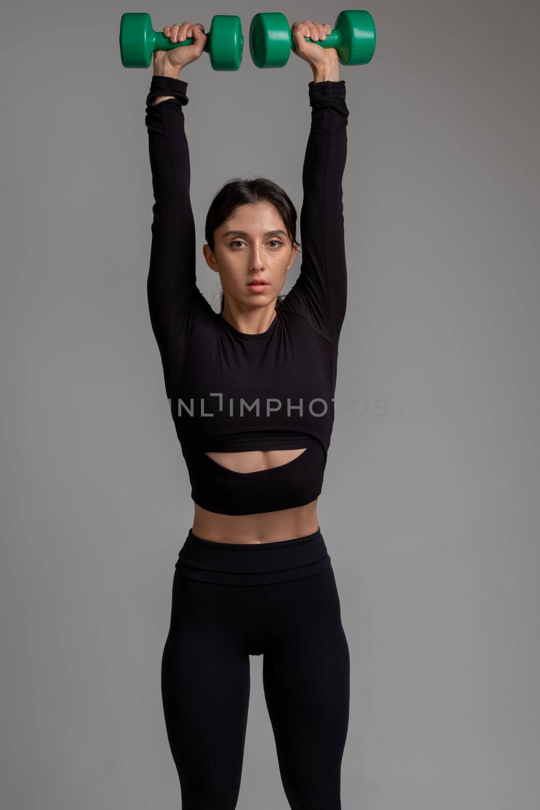 Concentrated fit brunette in black sportswear performing upper body workout with dumbbells on grey background. Set of exercises for shoulders. Fitness and bodybuilding concept
