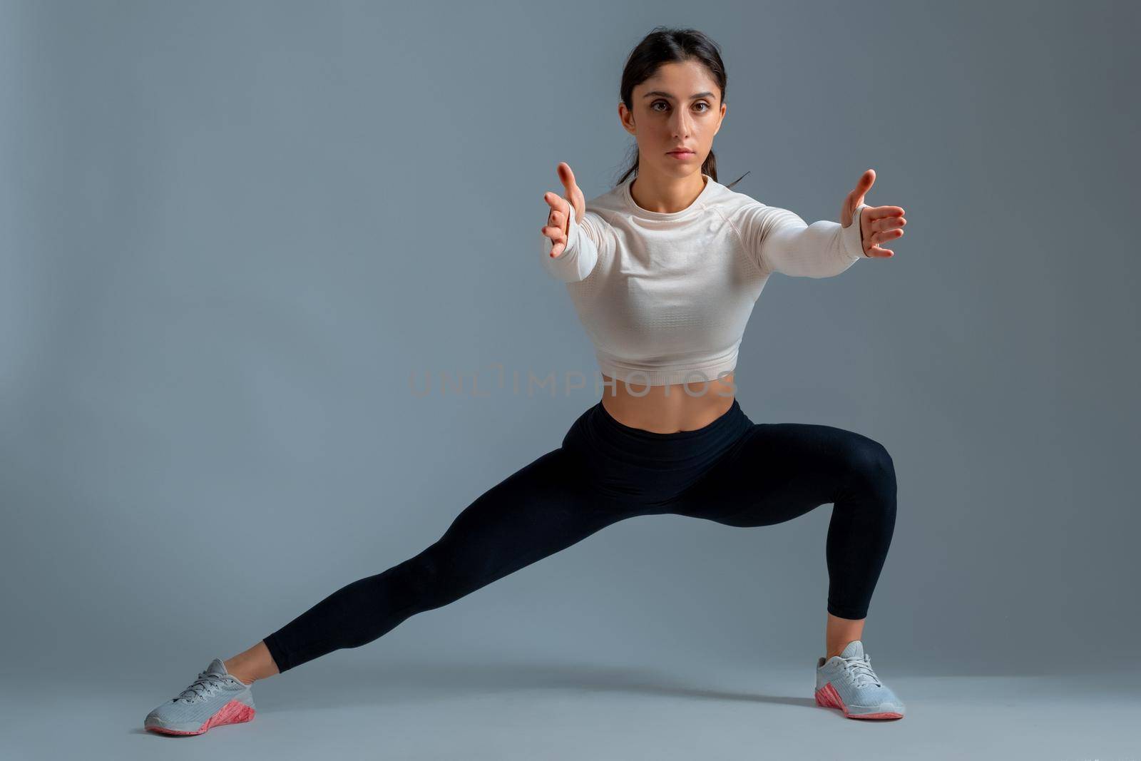 Sporty girl working out muscles of legs and glutes, doing bodyweight lateral split squats in studio on grey background. Fitness and sports motivation concept