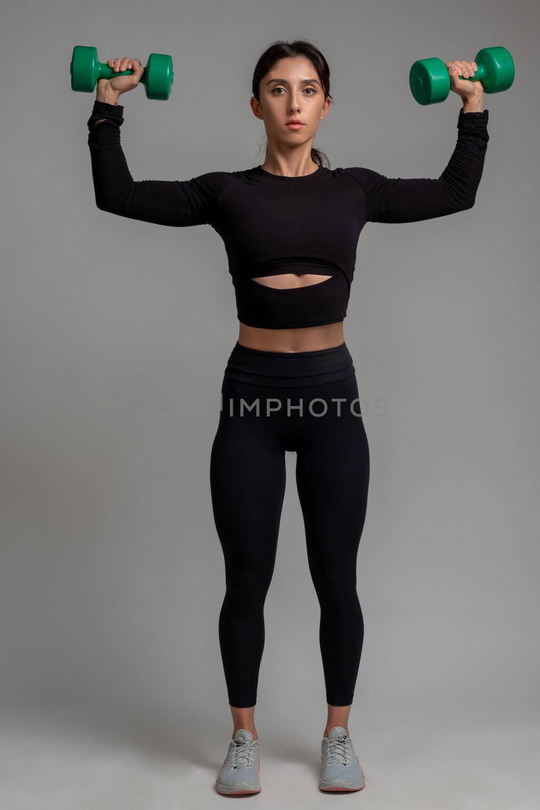 Focused fit brunette in black sportswear performing upper body workout in studio on grey background, doing dumbbell shoulder press. Fitness and bodybuilding concept