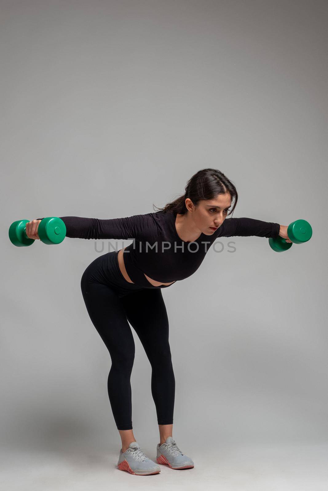 Athletic girl in black sportswear doing exercise to sculpt and tone shoulders, performing reverse fly with dumbbells on grey background. Sport and fitness concept