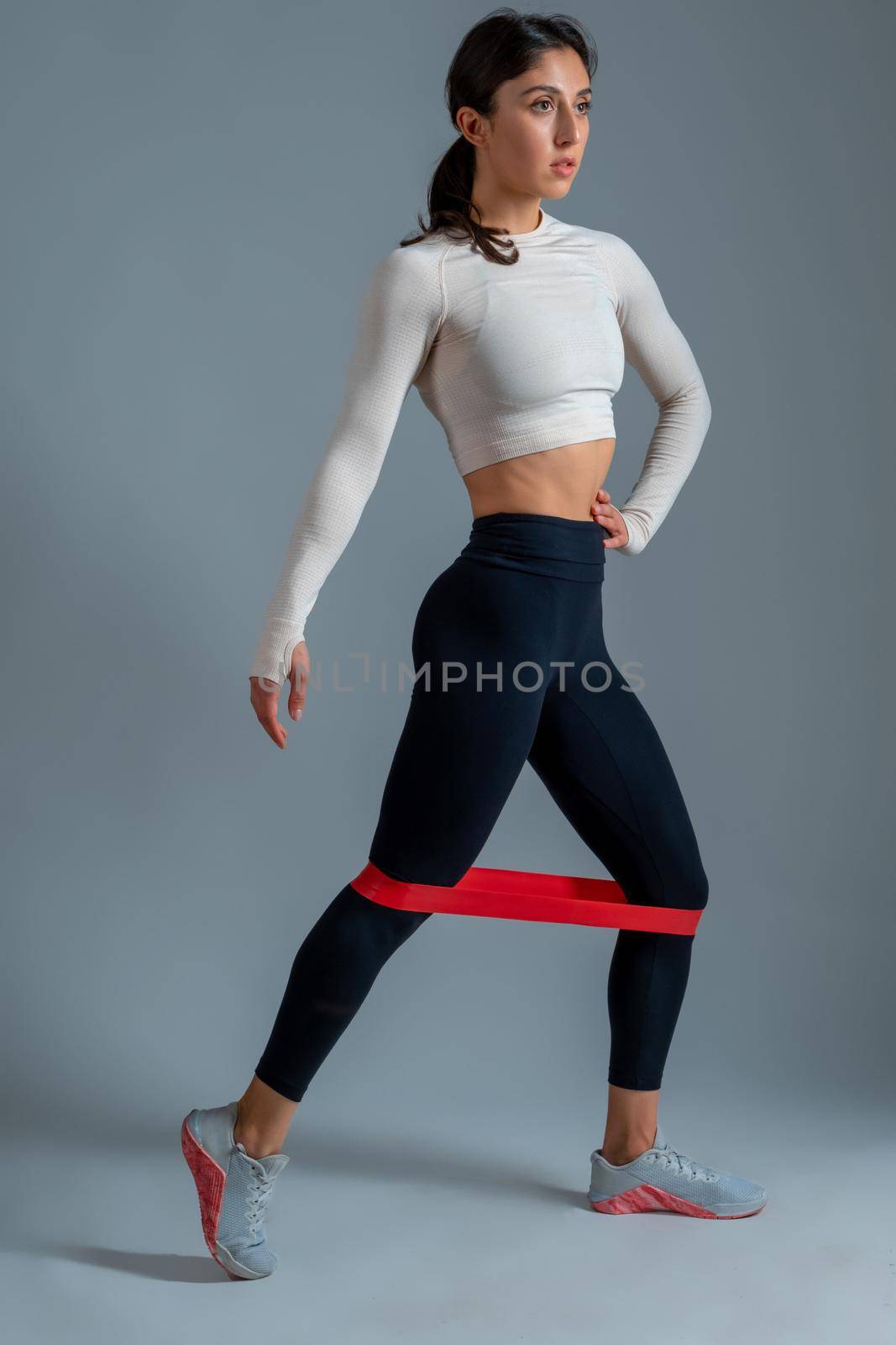 Full length portrait of athletic young woman performing leg and glute workout with loop resistance band in studio on grey background. Active lifestyle and fitness concept
