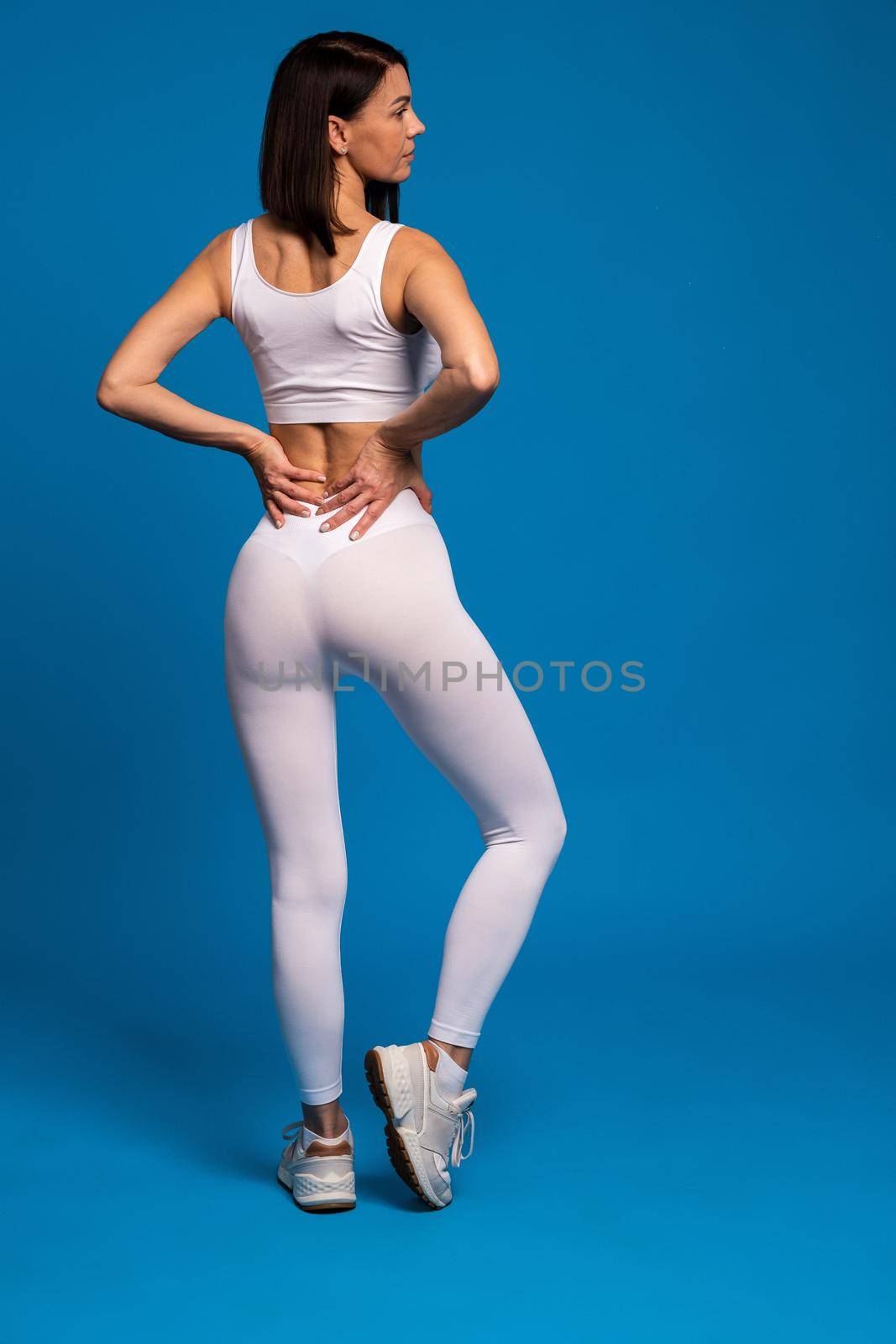 Rear view of fit brunette wearing white sports top and leggings demonstrating her good physical form on blue background. Healthy woman body as fitness results concept. Full length studio portrait