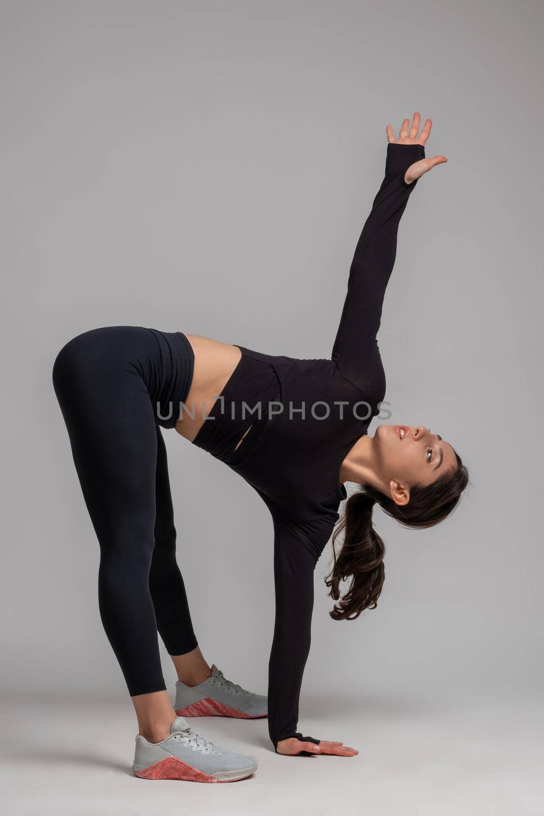 Young athletic brunette performing stretching workout to improve muscle elasticity and achieve comfortable muscle tone. Healthy lifestyle and fitness concept. Studio shot against grey background