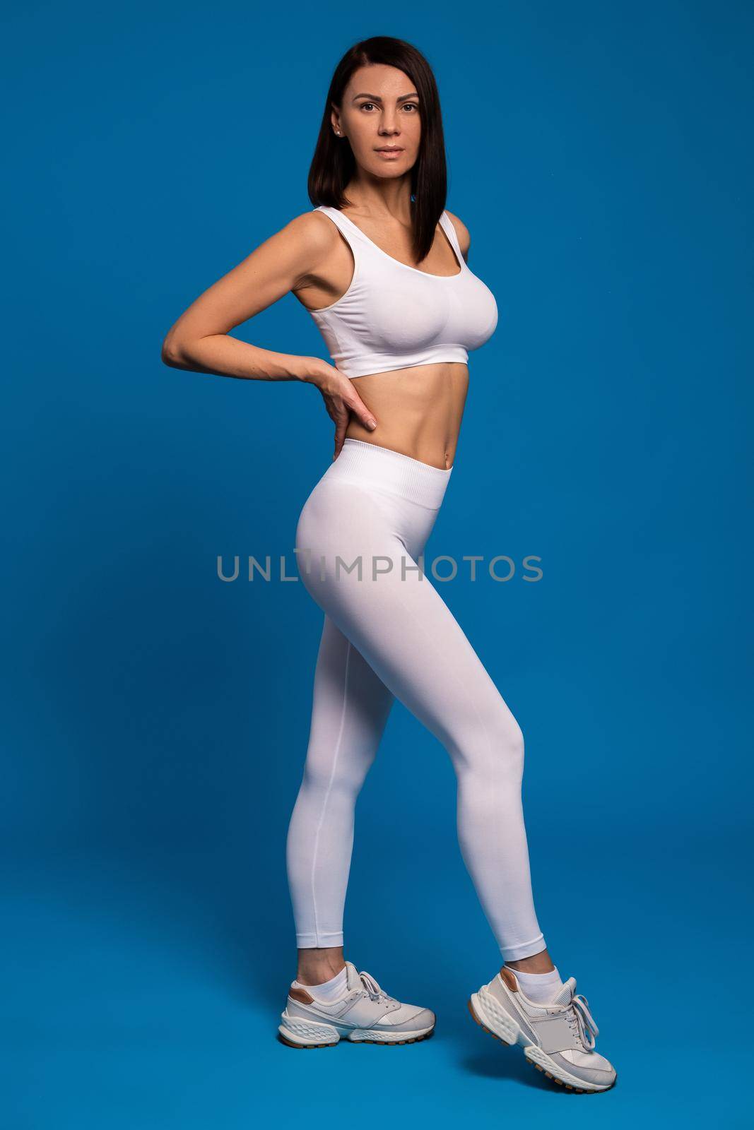 Full length portrait of real confident young brunette wearing white sports top and leggings demonstrating her good physical form on blue background. Healthy woman body as fitness results concept