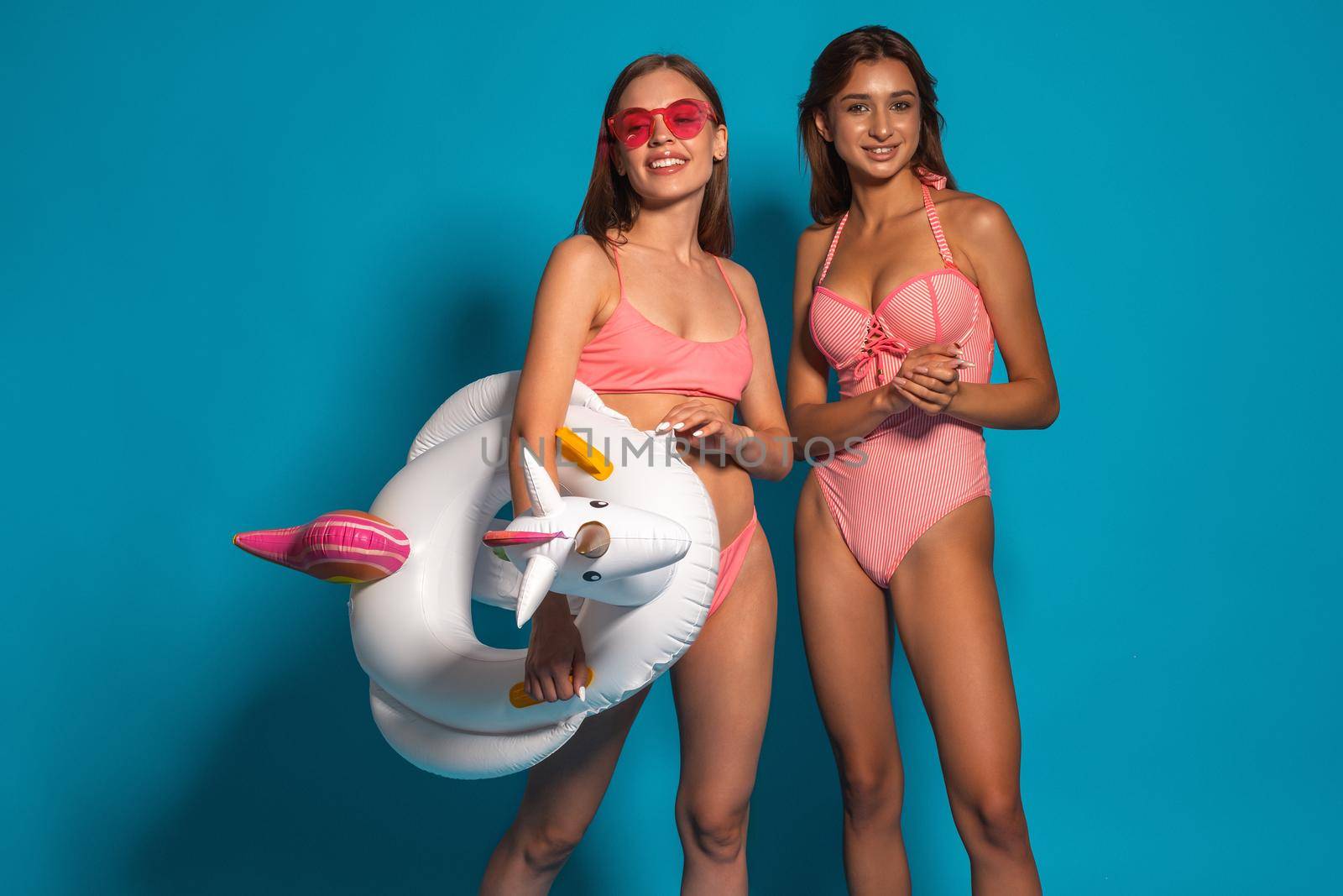 Two attractive smiling satisfied girls wearing bathing suits standing on blue studio background with rubber ring in form of funny unicorn. Beach holiday fun and enjoyment concept