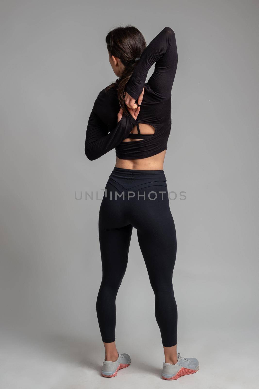 Rear view of sporty flexible girl doing stretching workout, standing with fingers clasped behind her back. Sportswoman training in studio on grey background