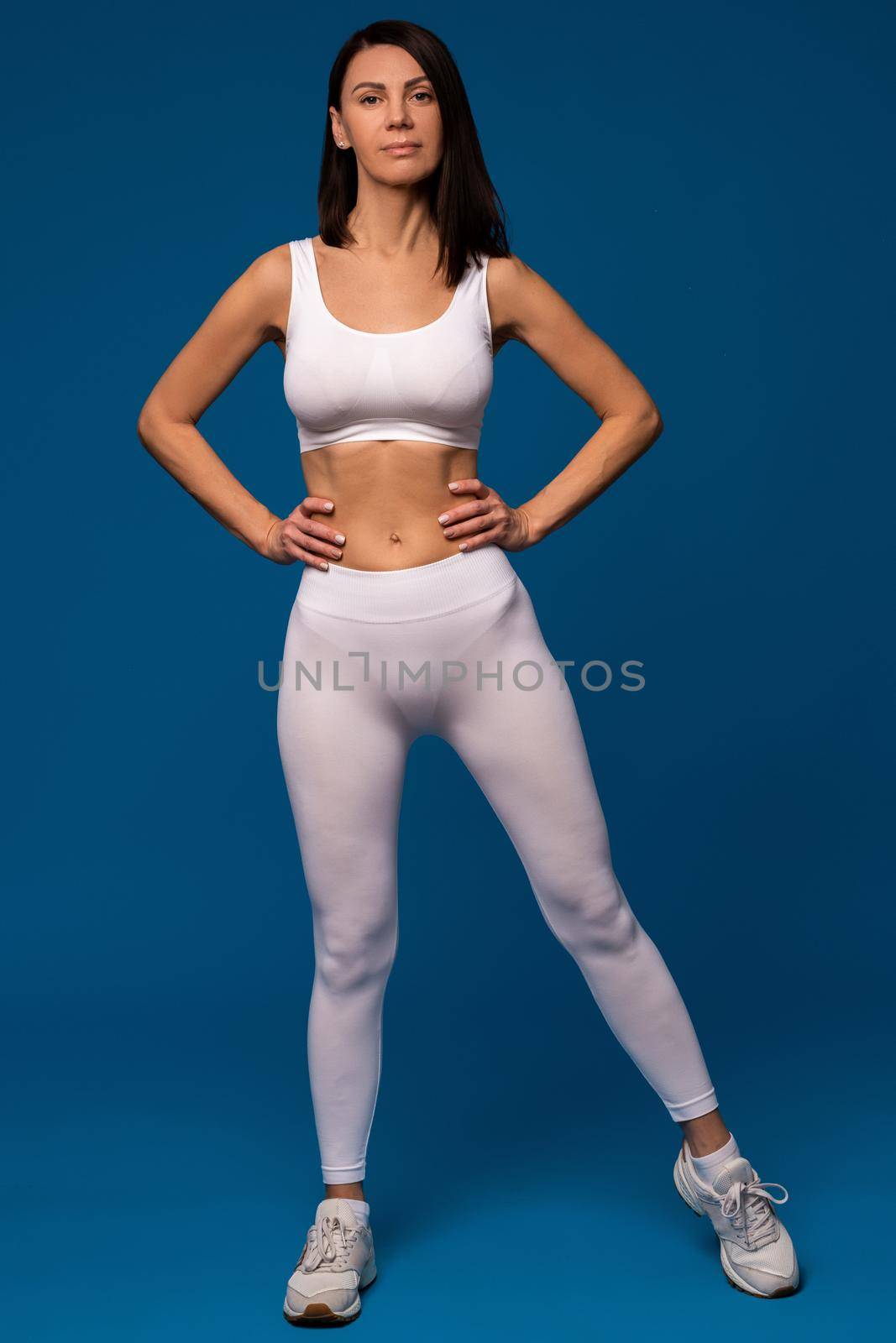 Portrait of fit sporty woman confidently posing in white activewear on blue background, proud of good her good physical shape. Concept of exercising and training results