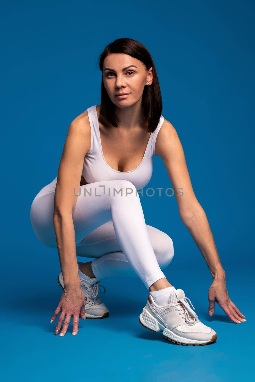 Confident fit woman wearing sports top and leggings posing during workout in studio, sitting on hunkers on blue background. Active lifestyle and fitness concept