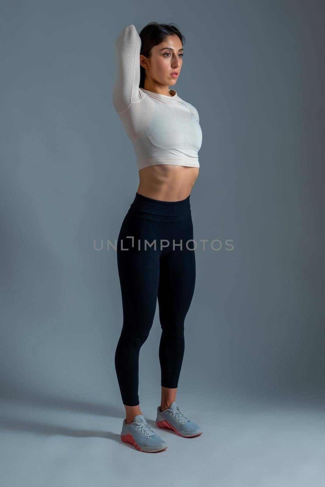 Focused slim girl doing stretching workout, standing with fingers clasped behind her back. Sportswoman training in studio on grey background