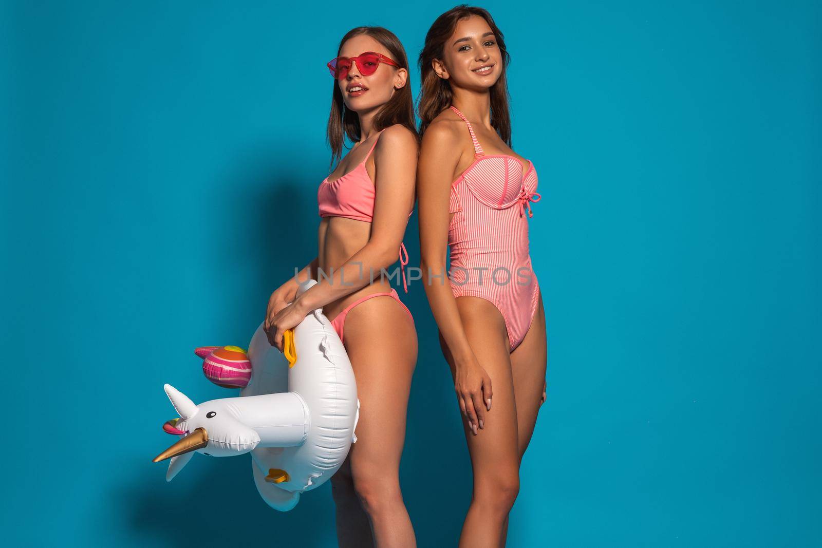 Two happy smiling girls in pink swimsuits standing back to back in studio on blue background, holding unicorn rubber ring. Concept of pool party or beach vacation