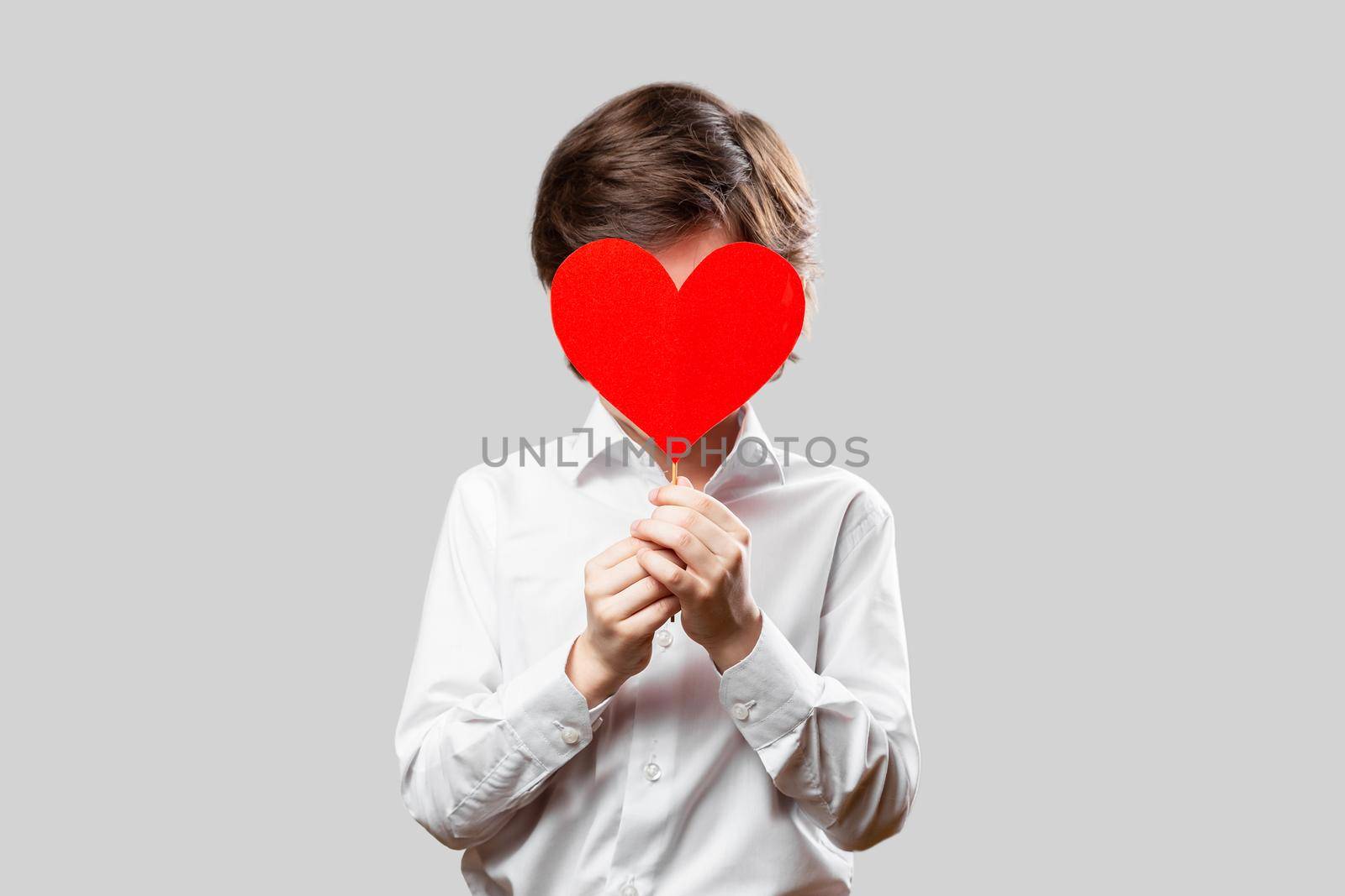 Boy hiding his face behind red st. valentines heart. Stylish young man wearing white shirt over grey background. Saint Valentines Day holiday concept